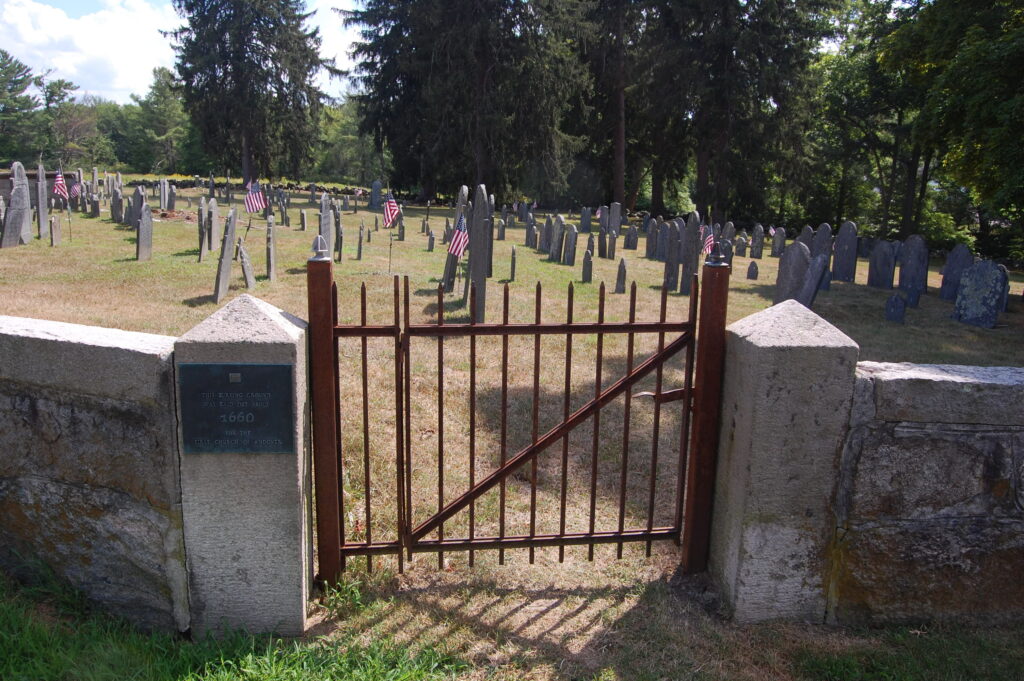 A rusty gate in a stone wall that enters into a cemetery with rows of gravestones in the background